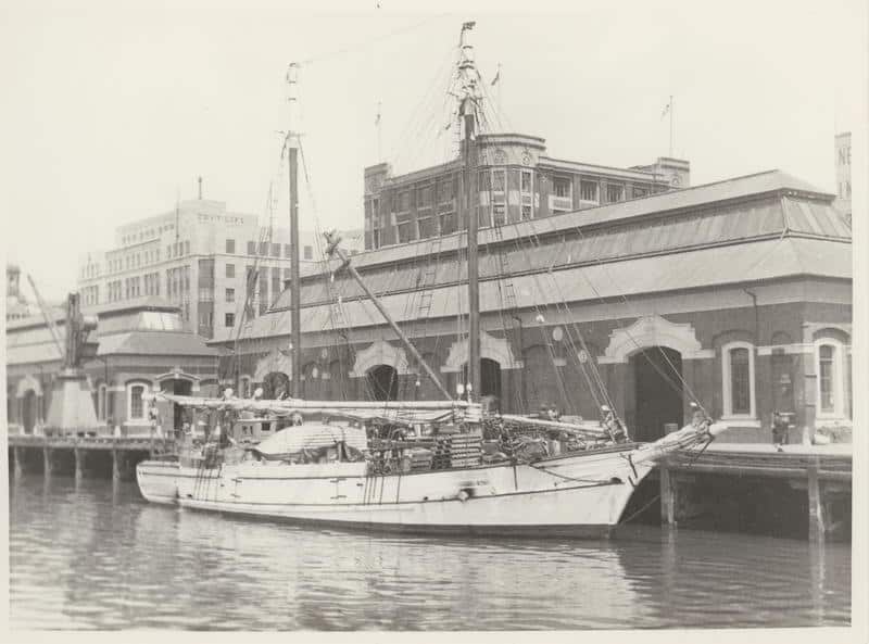 Black and white image of a scow called Kohi in 1911.  The boat is in front of a wharf with a building in the background.