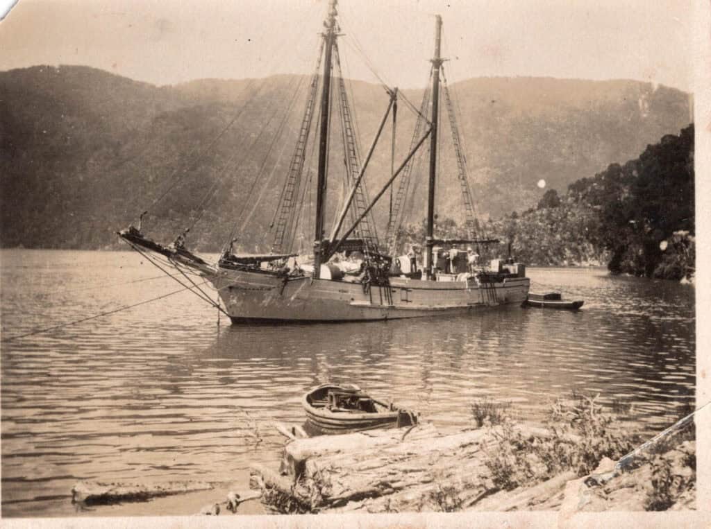 Sepia toned image of teh scow Kohi at anchor in Great Barrier Island. A dinghy tied to a log is in the foreground.