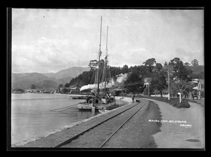 View of Haven Road Nelson with railway tracks on the waters edge and the scow Orakei tied to the side of the road.