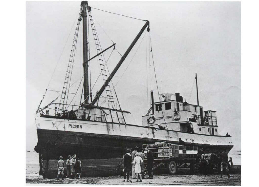 Black and white image of the side of Picton on the beach. Several people are standing in front of the boat as well as a trailer with several boxes is parked next to the boat