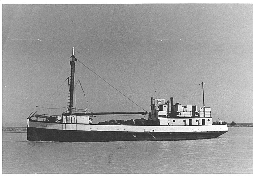 Black and white image of the side of Picton at sea. The deck is loaded and a person is sitting on the side.