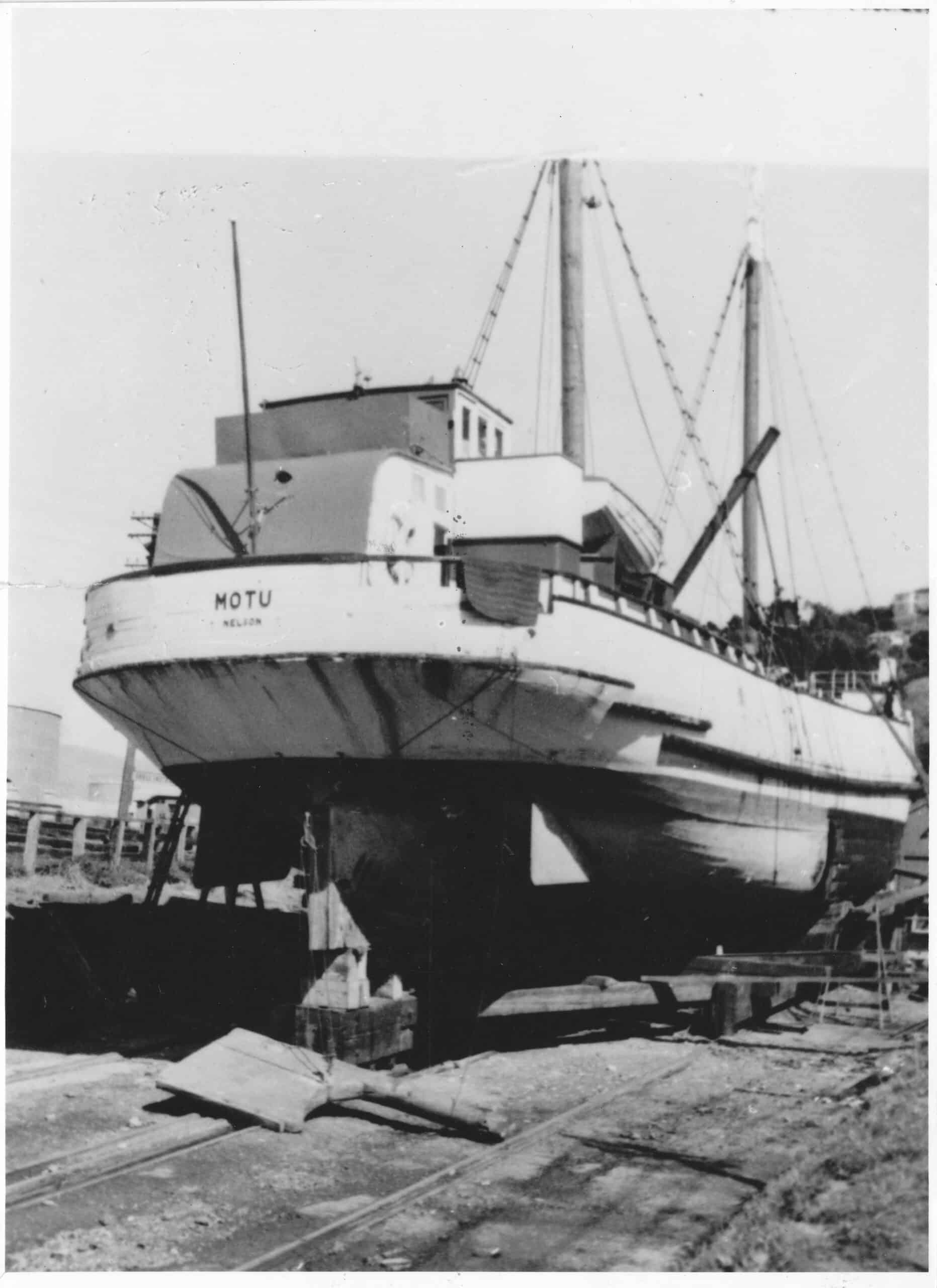 Black and white image of the scow Motu on the hard. The image is taken from the ground looking up at the boat with the slipway lines below and her rudder is on the ground