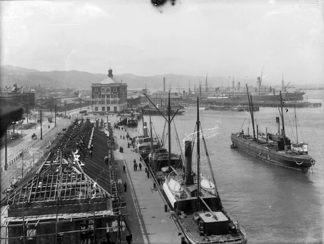 Black and white image of the Harbour Board Shed in Wellington under construction. Boats are tied to the wharf next to the shed and another is anchored in the water to the side.