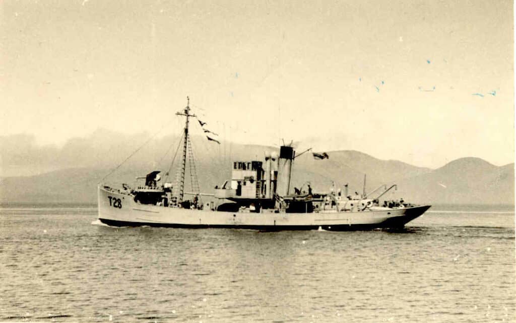 Black and white image of the minesweeper Pahau at sea. Seen from the side the boat has its T28 number clearly seen and flags flying.