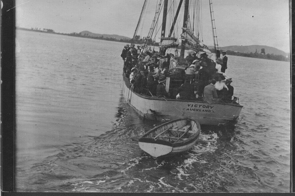 Black and white image of the scow Victory seen from the stern as she travels through the water. The decks are packed with people and a dinghy is towed behind. Small hills can be seen in the background.