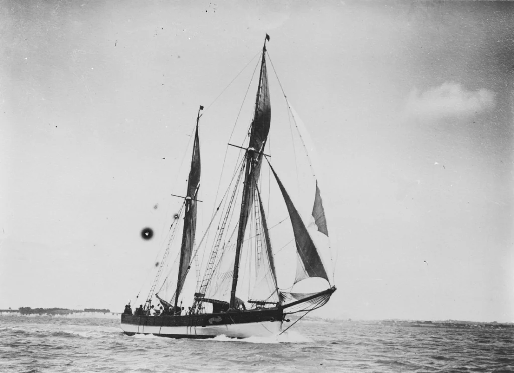 Black and white image of the scow Vixen seen from the front. She is under sail and has several people standing on the decks.